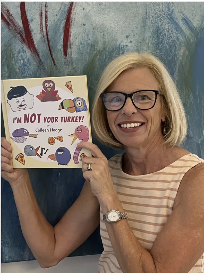 A smiling woman with blonde hair and glasses holds up a childrens book titled IM NOT YOUR TURKEY! by Colleen Hodge. The book cover features illustrated turkeys and food items.