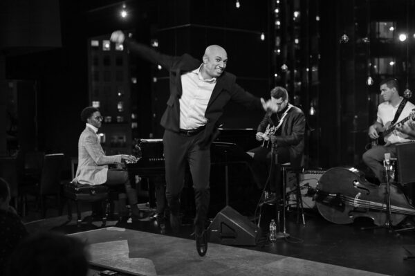 A man in a suit energetically dances on stage while a jazz band with a pianist, guitarist, and bassist performs behind him in a dimly lit venue.