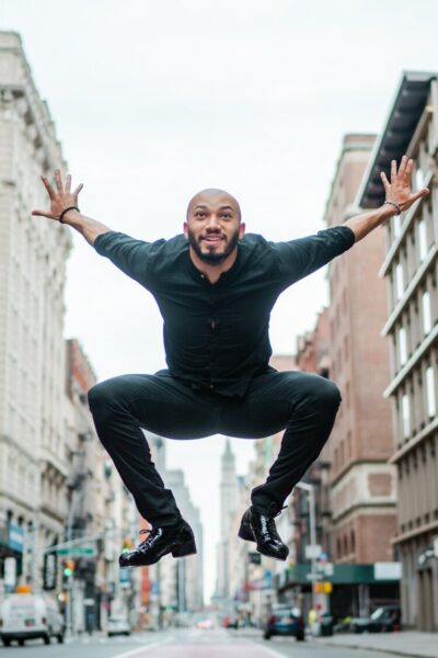 A man in a black outfit jumps mid-air on a city street, arms spread wide and smiling, with tall buildings lining both sides of the street and cars visible in the background.