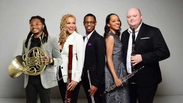 Five musicians in formal attire stand smiling with their instruments, including a French horn, bassoon, clarinet, flute, and oboe, against a plain light gray background.