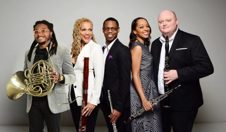 Five musicians in formal attire stand smiling with their instruments, including a French horn, bassoon, clarinet, flute, and oboe, against a plain light gray background.