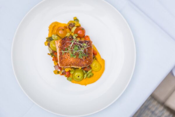 A plated dish featuring a seared salmon fillet on top of a colorful vegetable medley and a bright orange sauce, garnished with microgreens, served on a white plate.