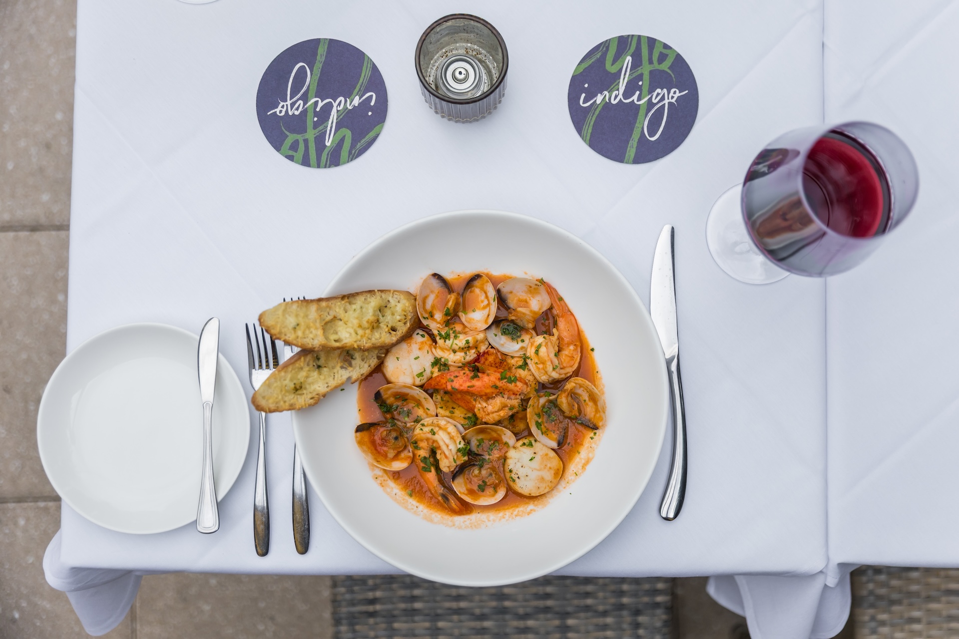 A plate of seafood pasta with clams, shrimp, and scallops in tomato sauce, garnished with herbs and served with toasted bread. A glass of red wine and utensils are arranged on a white tablecloth.