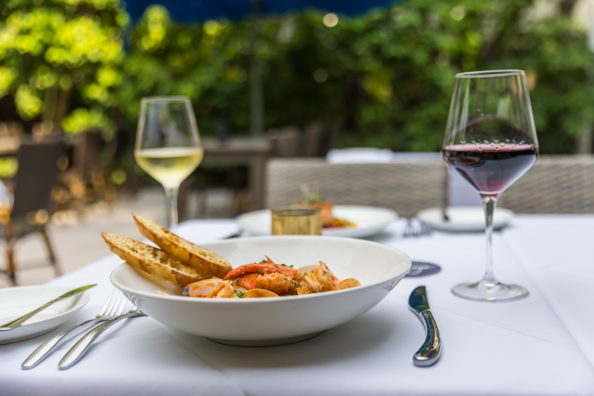 A white bowl filled with shrimp and toasted bread sits on a table set for two, alongside glasses of white and red wine, with a leafy outdoor background.