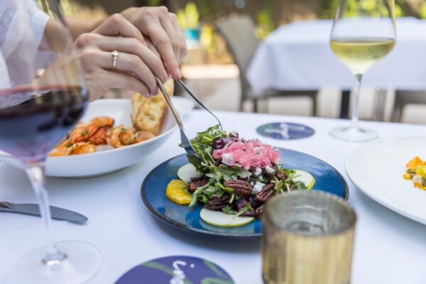 A person uses a fork and knife to eat a fresh salad with pecans and pickled onions on a blue plate, with wine glasses and other dishes on a white tablecloth in an outdoor dining setting.