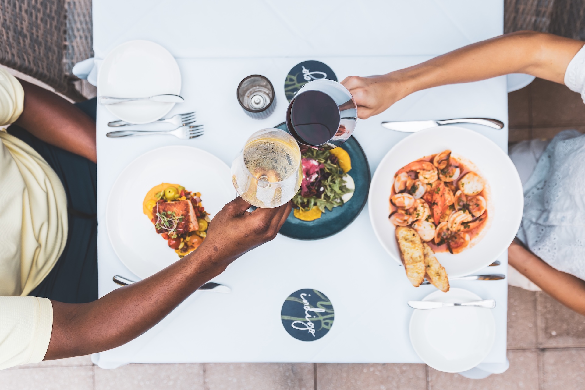 Two people clink wine glasses over a table set with plates of seafood, salad, and bread; the scene is viewed from above, showing a relaxed, elegant dining atmosphere.