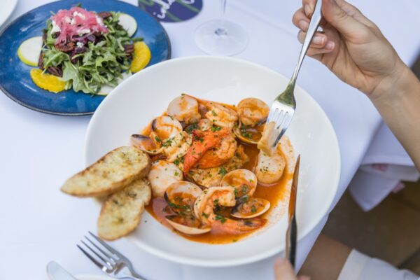 A person cuts into seafood in a white bowl with shrimp, clams, scallops, and tomato sauce, served with toasted bread; a plate of salad with orange slices is nearby on a white tablecloth.