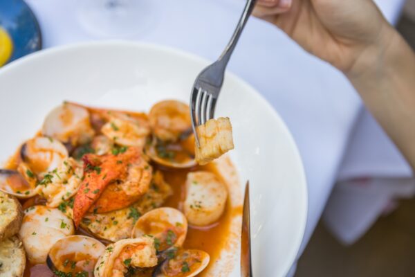 A hand holding a fork with a piece of seafood above a bowl of seafood stew at Indigo restaurant on Amelia Island, Florida, containing shrimp, clams, lobster, bread, and herbs in a tomato-based broth