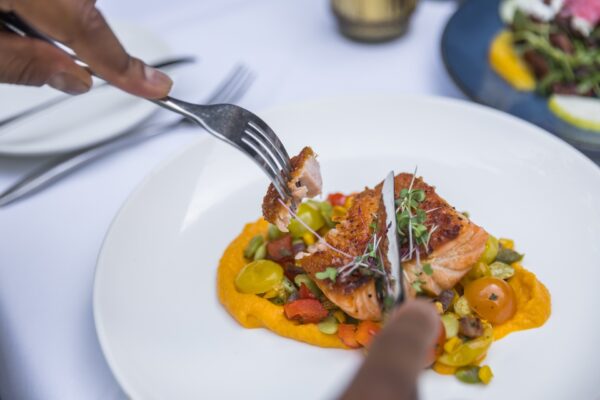 A person cutting into a piece of seared salmon at Indigo restaurant on Amelia Island, Florida, served on a bed of colorful vegetables and orange puree on a white plate.