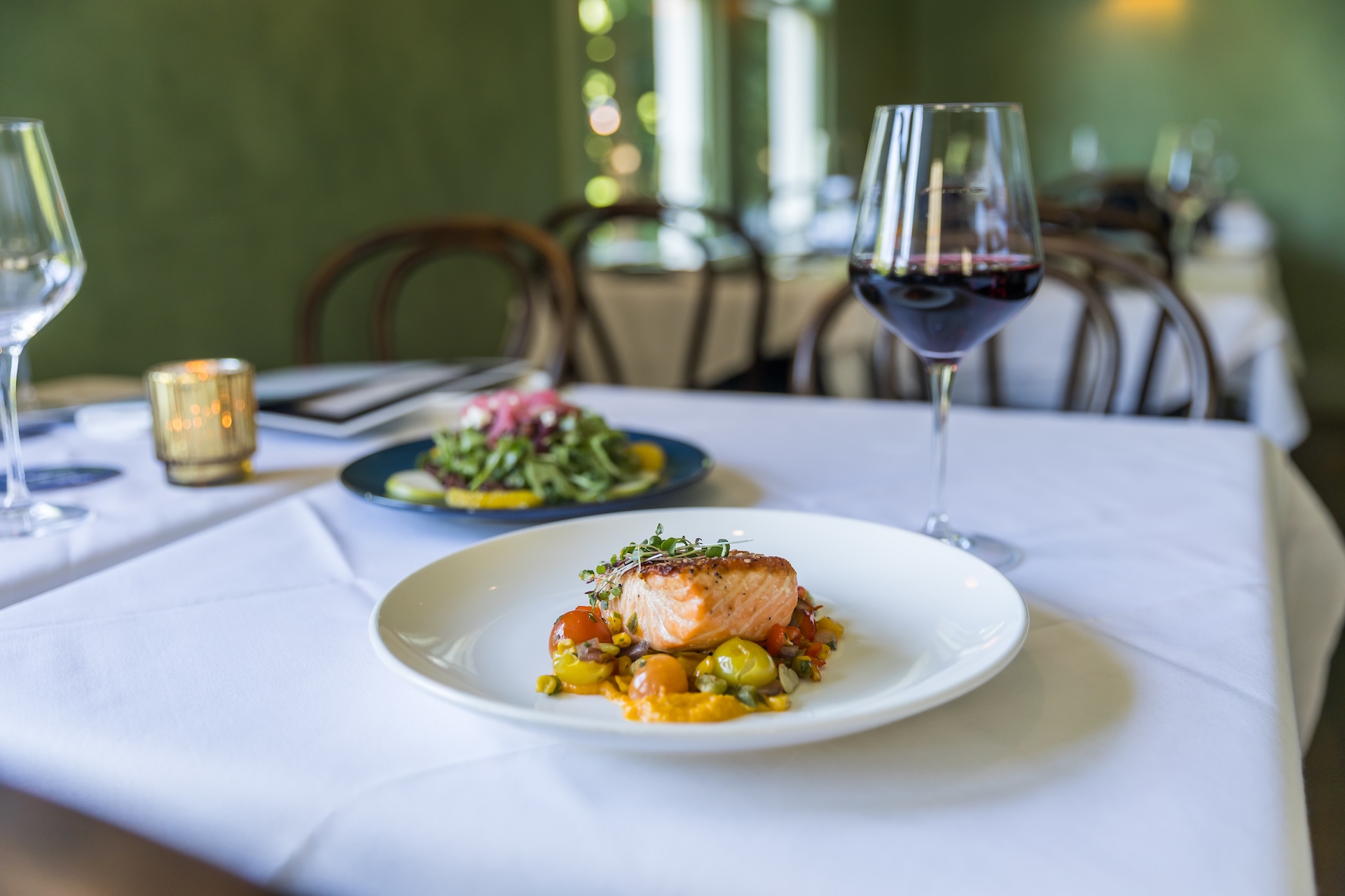A white plate with grilled salmon on a bed of cherry tomatoes and herbs sits on a white tablecloth. Next to it is a glass of red wine and another plate of salad in a softly lit, elegant restaurant.