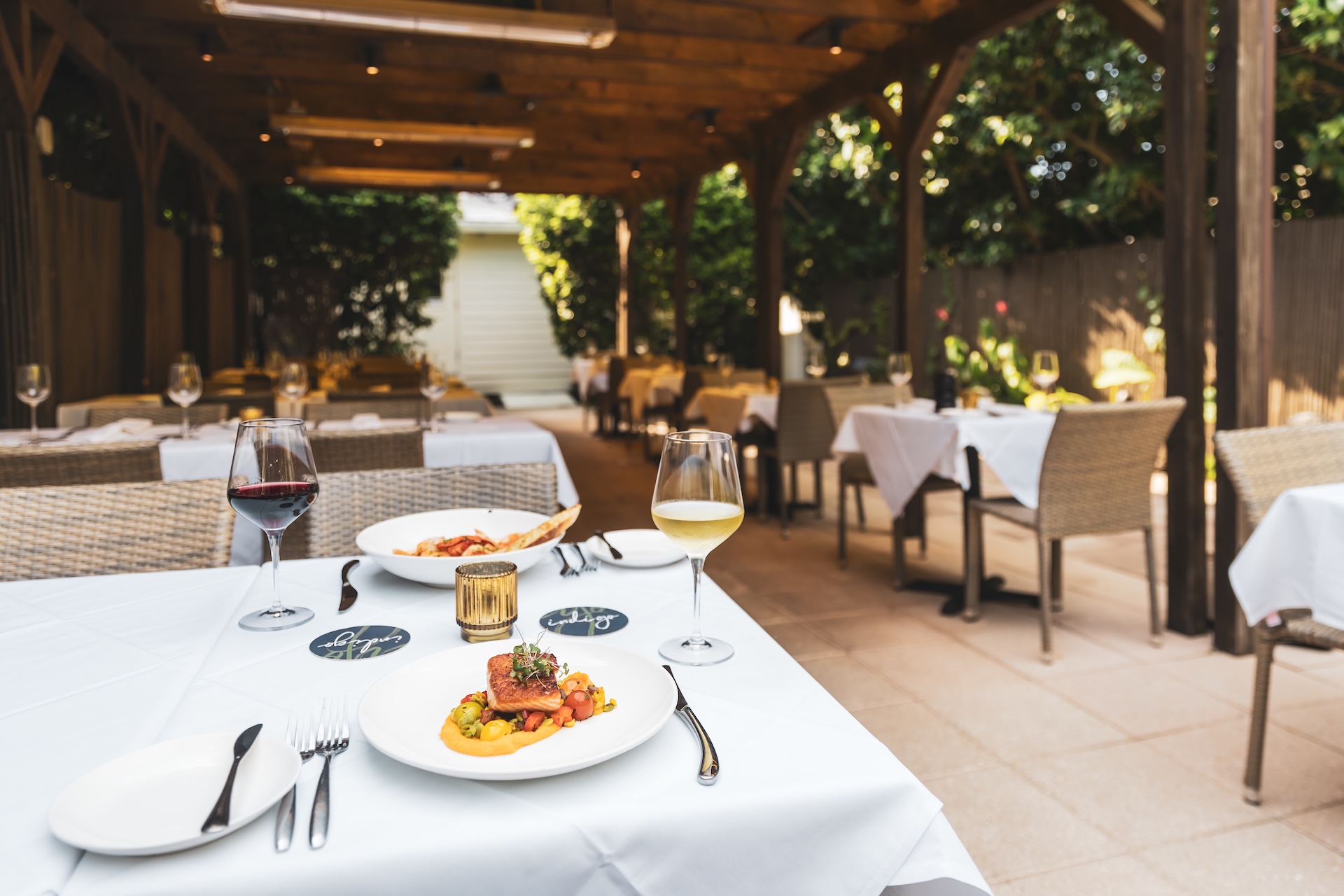 A covered outdoor restaurant patio with white tablecloths, neatly set tables, wine glasses, and gourmet food. Sunlight streams through the wooden pergola, and greenery surrounds the cozy dining area.