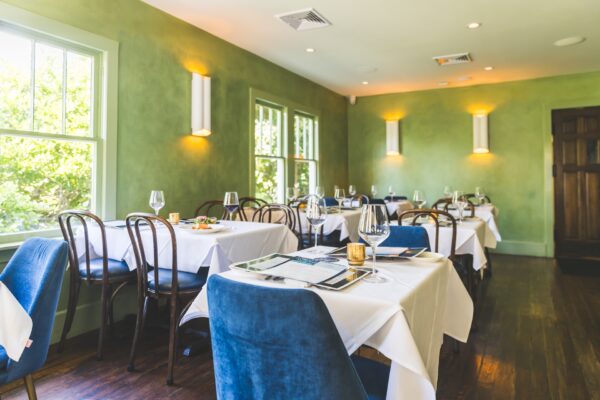 A bright, elegant restaurant dining room with green walls, wooden chairs, blue upholstered chairs, white tablecloths, and neatly set tables near large windows letting in natural light.
