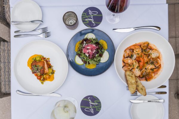 A top-down view of a table set with three dishes: a salad with greens and orange slices, a seafood pasta with bread, and a dish with fish and vegetables. Two glasses of wine and cutlery are also visible on a white tablecloth.