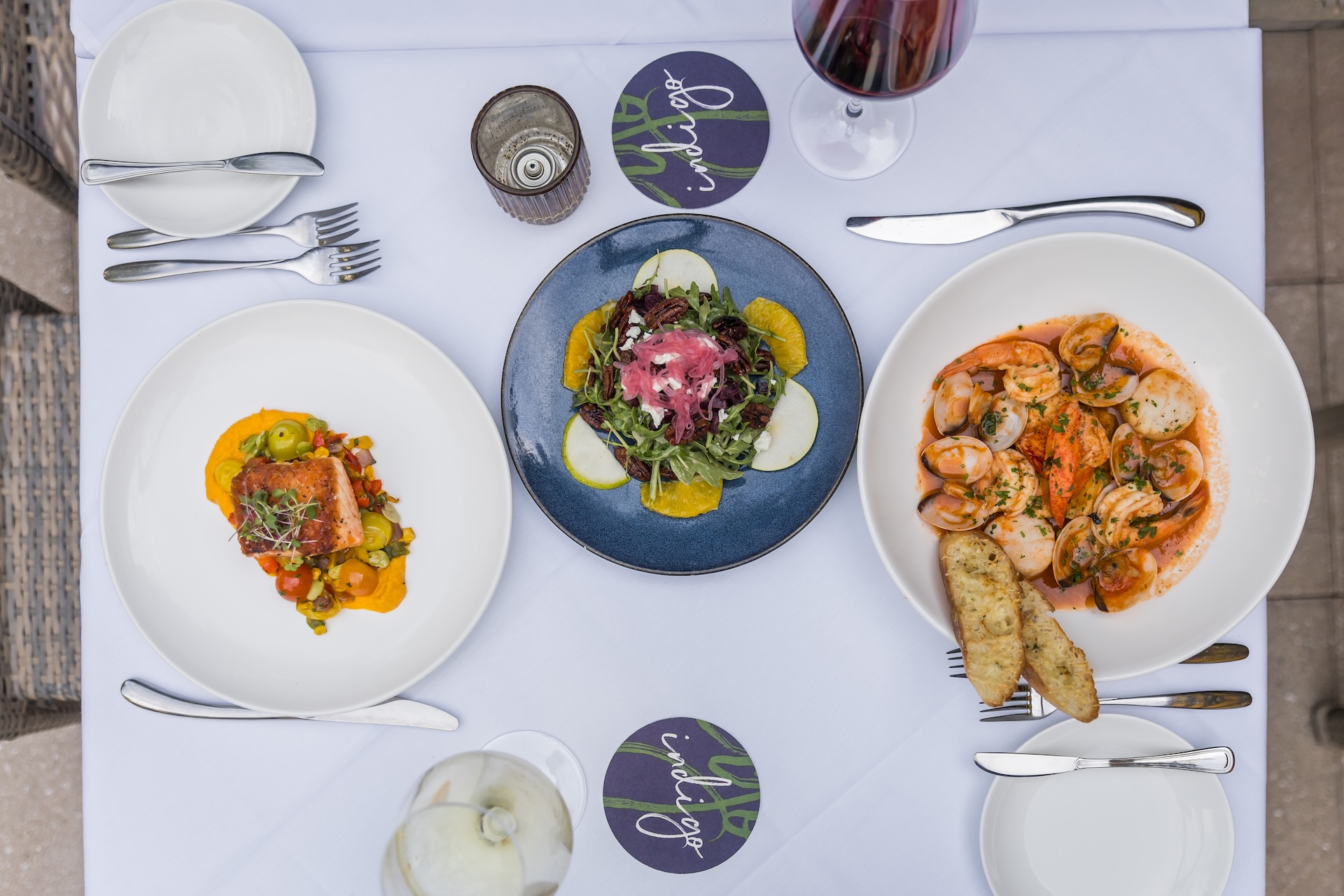 A top-down view of a table set with three dishes: a salad with greens and orange slices, a seafood pasta with bread, and a dish with fish and vegetables. Two glasses of wine and cutlery are also visible on a white tablecloth.