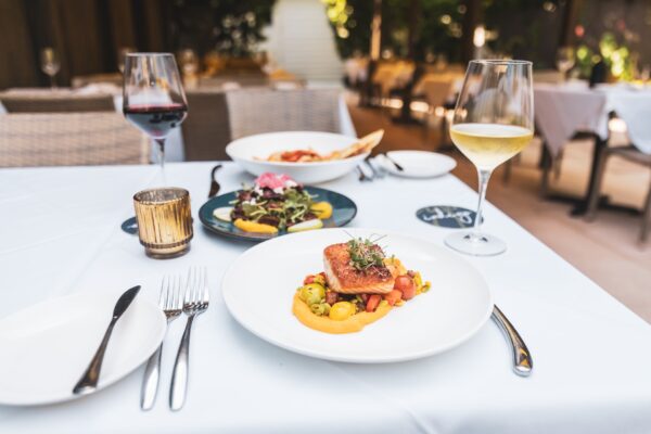 A white tablecloth dining table set with a plate of gourmet salmon, a salad, bread, red and white wine glasses, and a candle, in an elegant outdoor restaurant setting.
