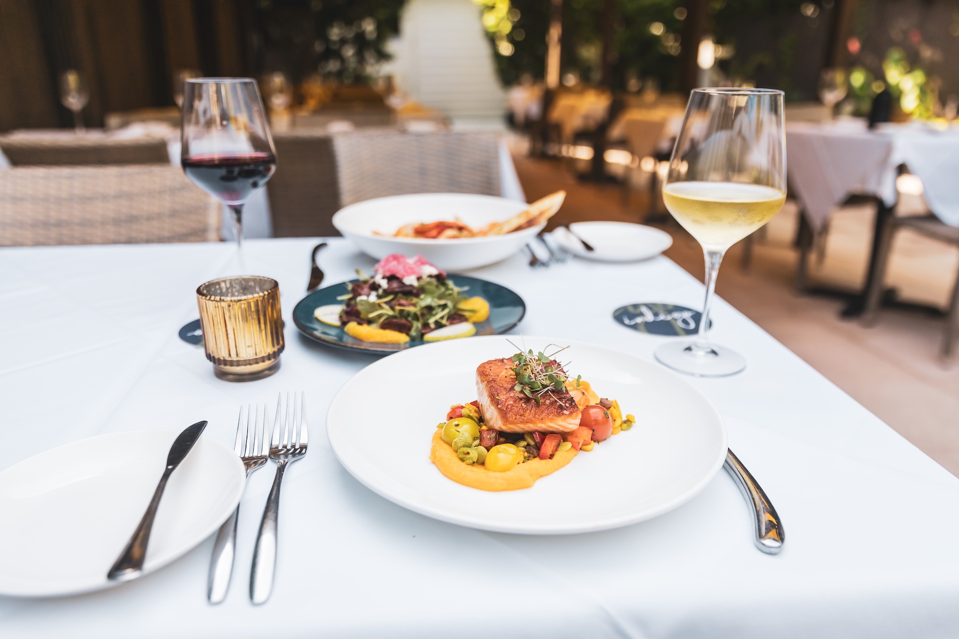 A white tablecloth dining table set with a plate of gourmet salmon, a salad, bread, red and white wine glasses, and a candle, in an elegant outdoor restaurant setting.