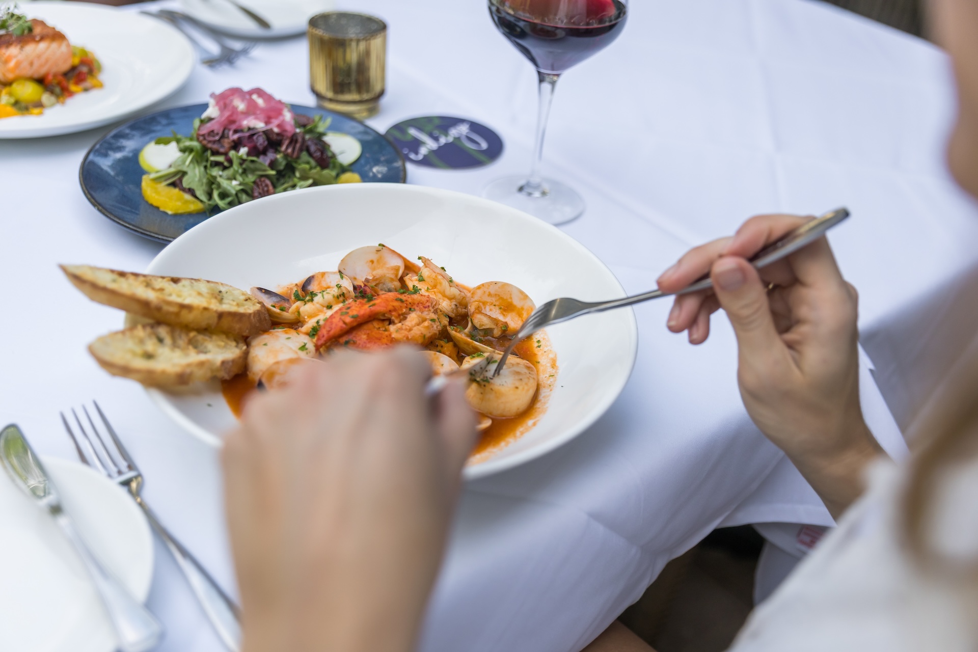 A person eating a seafood dish with shrimp and sauce, accompanied by toasted bread, at a white tablecloth restaurant table set with utensils, wine, and salad plates.