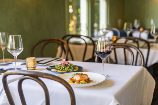 A white-clothed restaurant table is set with a plate of salmon and vegetables, a glass of red wine, empty wine glasses, and a lit candle. More tables and chairs are visible in the softly lit background.