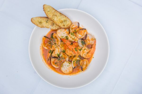 A white bowl of seafood stew with clams, shrimp, and scallops in a tomato-based broth, garnished with herbs, served with two slices of toasted garlic bread on a white tablecloth.
