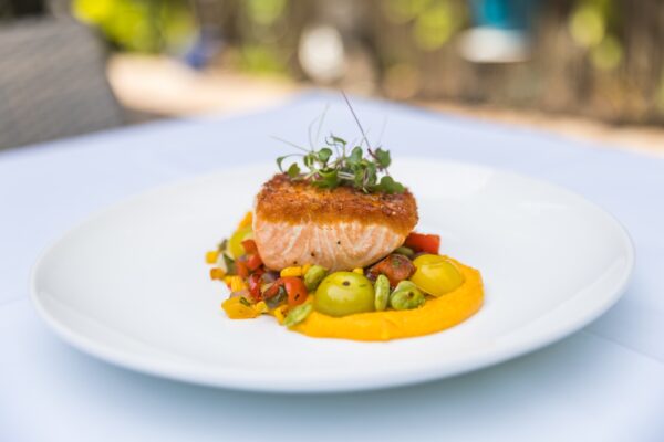 A seared salmon fillet garnished with microgreens sits atop a bed of colorful diced vegetables and a bright orange puree on a white plate, with a blurred outdoor background.