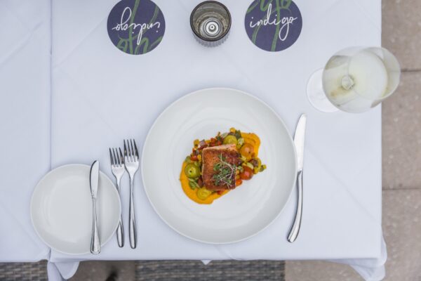 A white tablecloth with a plate of seared fish atop colorful vegetables and a sauce, next to a fork, knife, and spoon, a glass of white wine, two branded coasters, and a bread plate with utensils.