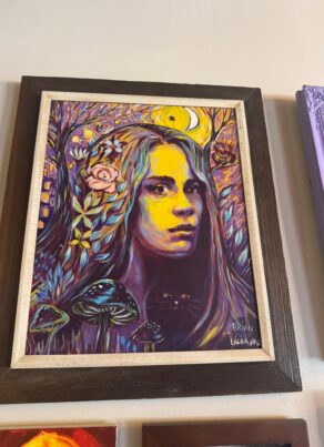 A vibrant painting of a woman’s face with long hair, surrounded by flowers, mushrooms, and trees in bold purples, blues, and yellows. A crescent moon and small houses are in the colorful, mystical background.
