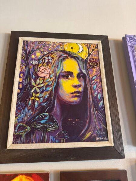A vibrant painting of a woman’s face with long hair, surrounded by flowers, mushrooms, and trees in bold purples, blues, and yellows. A crescent moon and small houses are in the colorful, mystical background.