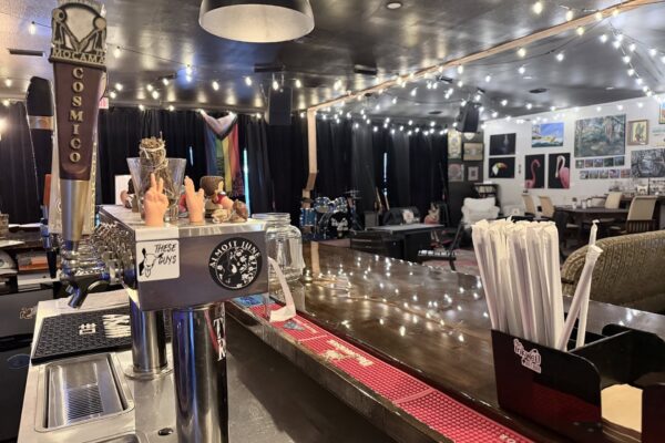 A cozy bar with beer taps, jars, and quirky decorations on the counter, string lights overhead, art on the walls, and a small stage with musical instruments in the background.