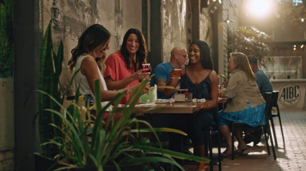 A group of people sit at outdoor tables along a rustic, weathered wall, enjoying drinks and conversation in a lively, sunlit setting, with plants in the foreground.