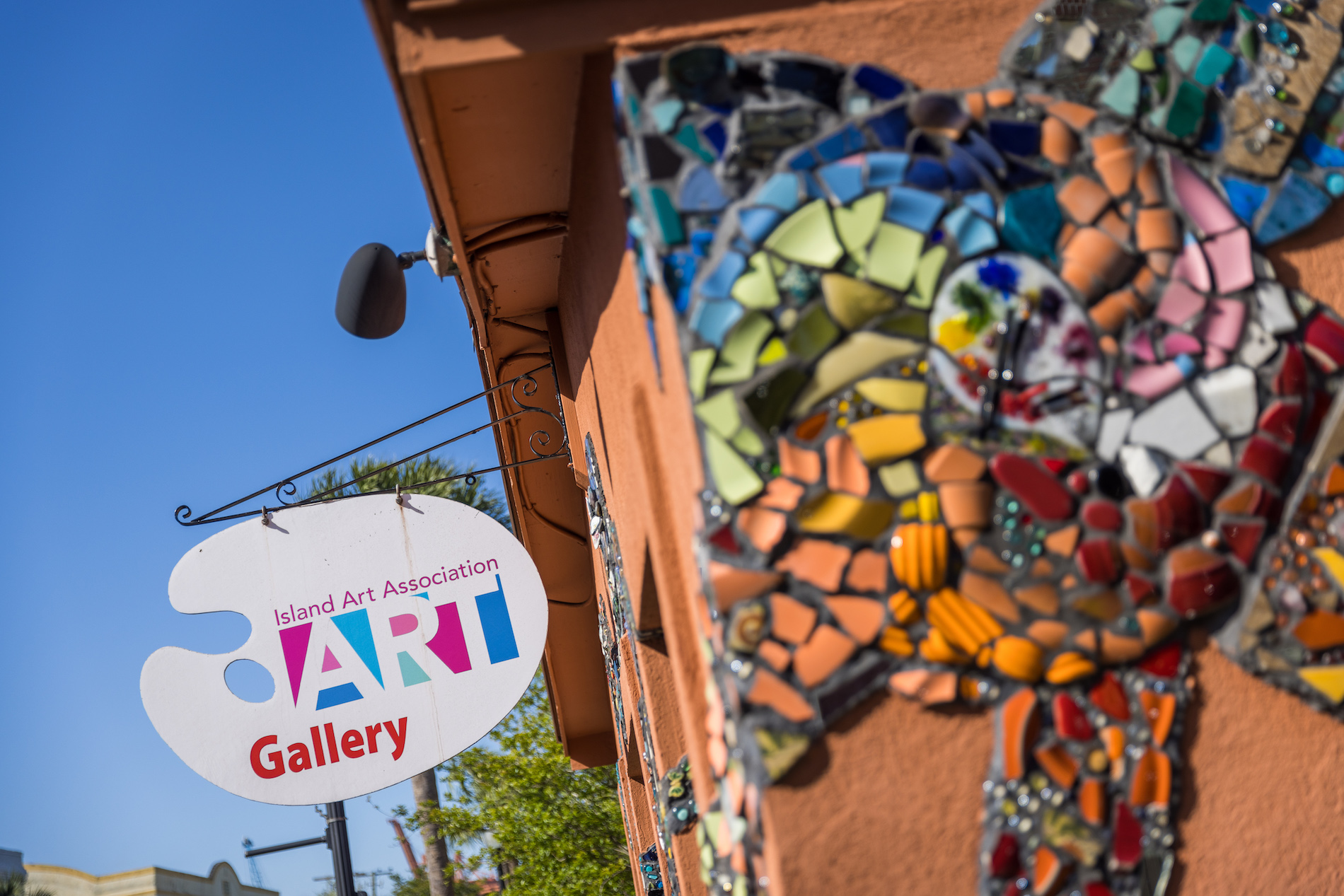 A close-up of a colorful mosaic on a building wall next to a hanging sign that reads “Island Art Association ART Gallery” under a clear blue sky.