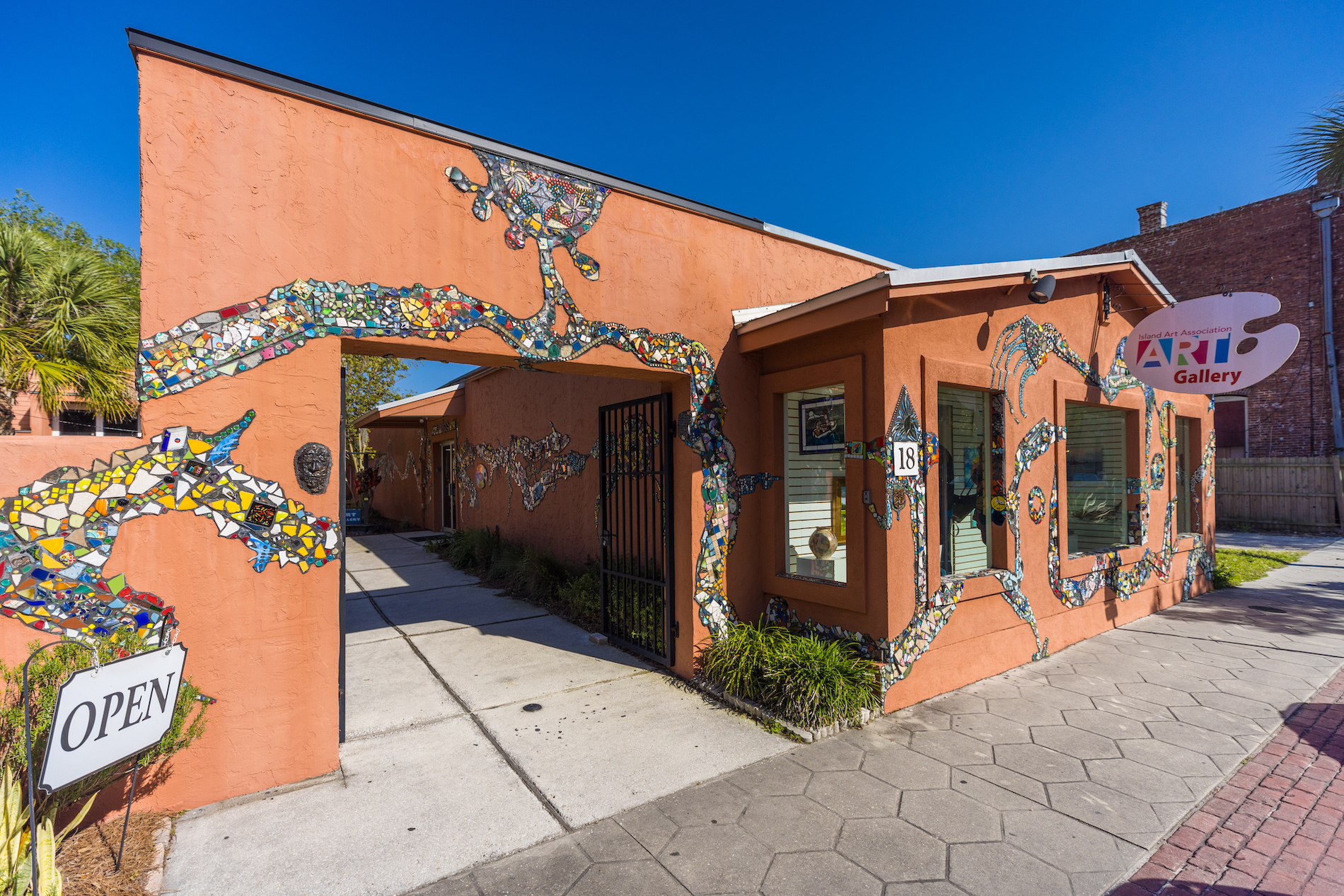 An orange art gallery building with a colorful mosaic design along the exterior wall. A sign reading OPEN is near the entrance, and a larger sign above says ART Gallery. The sky is clear and blue.