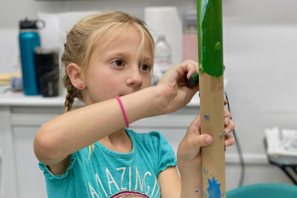 A young girl with blonde hair in braids paints a cardboard tube green. She wears a blue shirt with AMAZING on it and a pink bracelet, focusing intently on her work in a classroom or craft room setting.