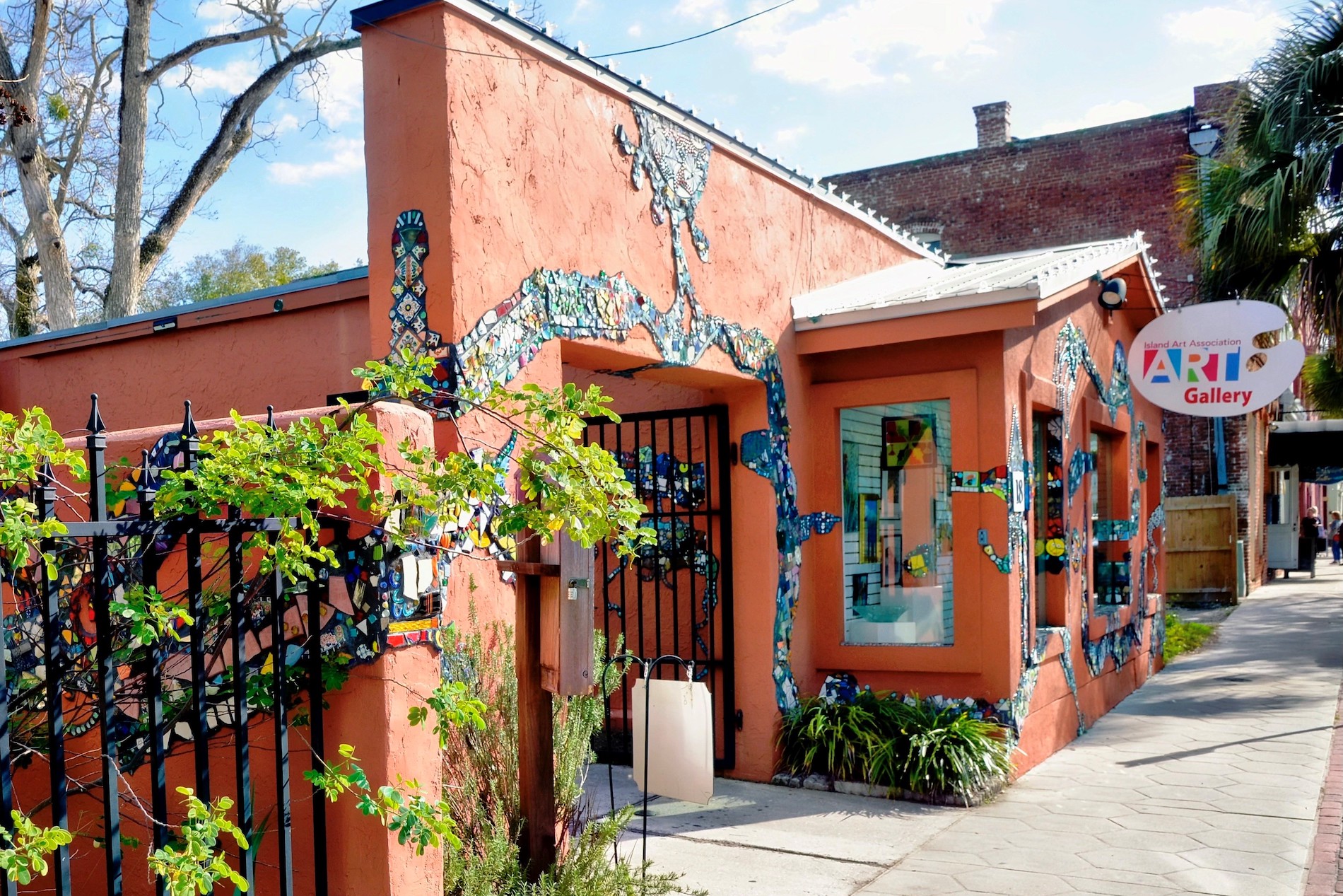 A small art gallery with orange walls decorated with colorful mosaic designs, a white “Art Gallery” sign, metal fence, greenery, and a sidewalk under a partly cloudy sky.