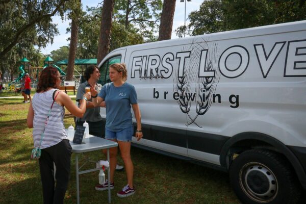 Three people stand by a van labeled First Love Brewing at an outdoor event. Two women behind a small table serve drinks to a guest in a park setting with trees and playground equipment visible in the background.