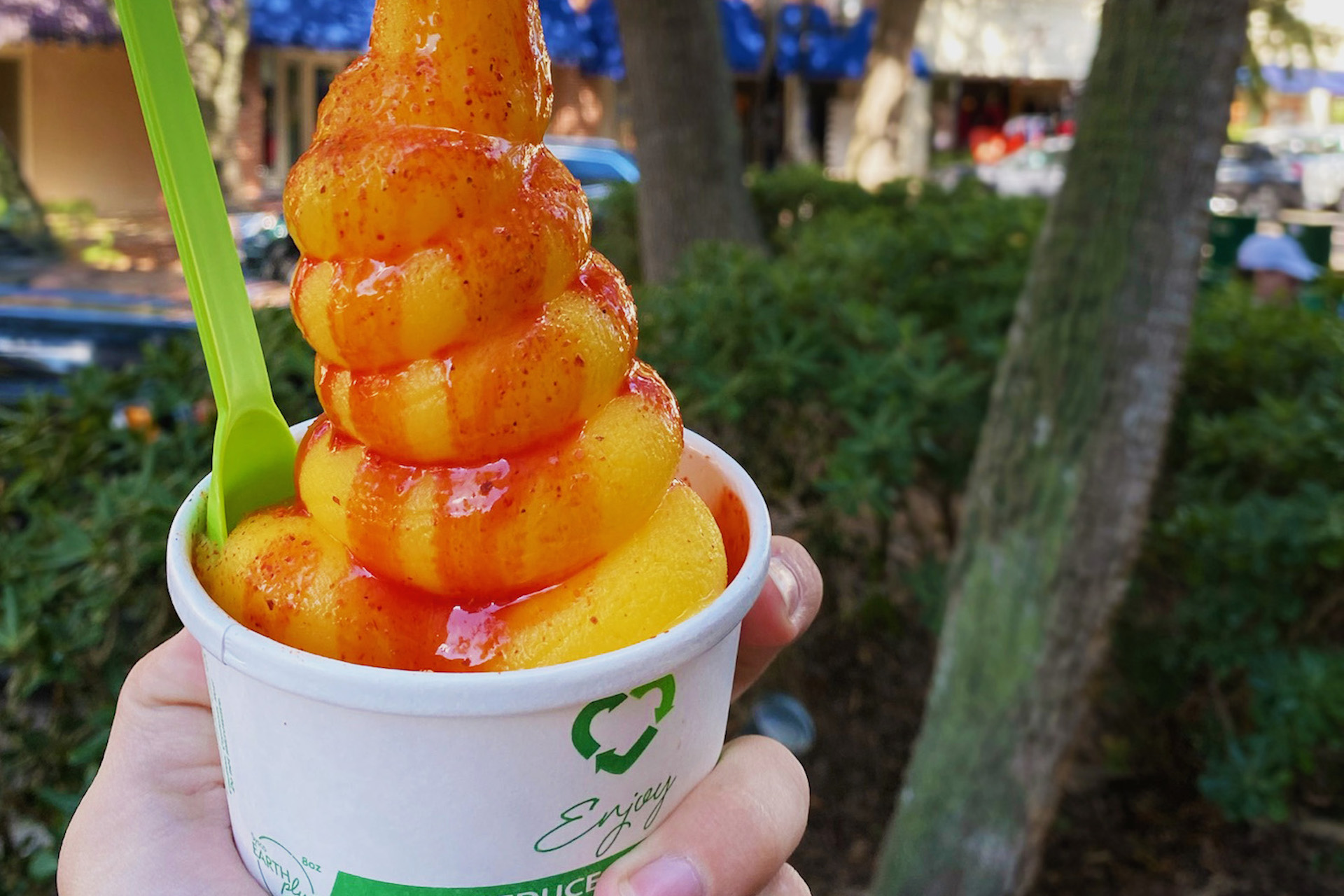 A hand holds a cup of mango sorbet topped with chili powder and chamoy sauce, with a green spoon beside it. Trees and an outdoor scene are blurred in the background.