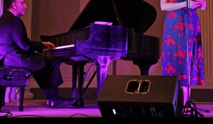 A man in a suit plays a grand piano while a woman in a floral dress sings into a microphone on a stage with purple lighting.