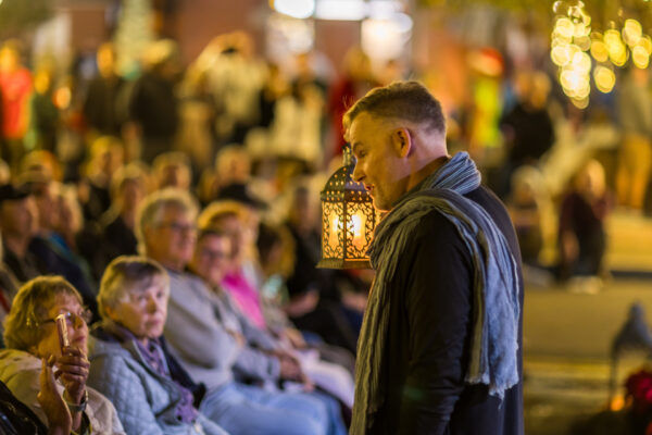 A man wearing a scarf stands and speaks to a seated audience outdoors at night. The area is warmly lit, and decorative string lights and a lantern hang nearby, while a blurred crowd is visible in the background.