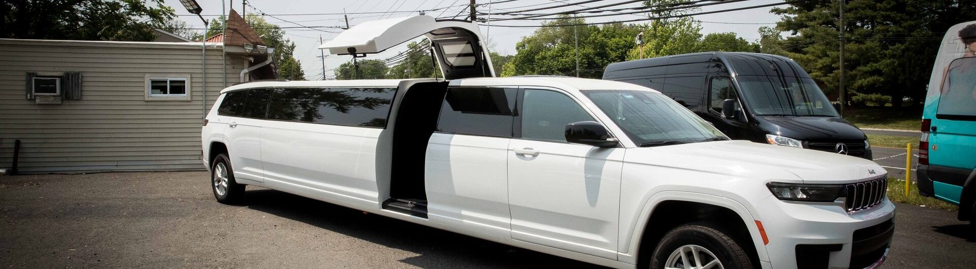 A long white stretch limousine SUV is parked outside with its gull-wing door open; other vans, power lines, and trees are visible in the background.