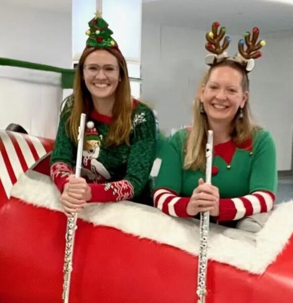 Two women wearing festive holiday sweaters and headbands—one with a Christmas tree, one with reindeer antlers—smile while holding flutes and sitting on a large red and white sleigh indoors.
