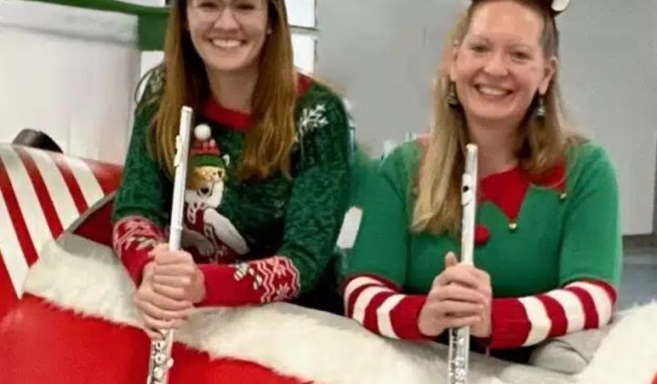 Two women wearing festive holiday sweaters and headbands—one with a Christmas tree, one with reindeer antlers—smile while holding flutes and sitting on a large red and white sleigh indoors.