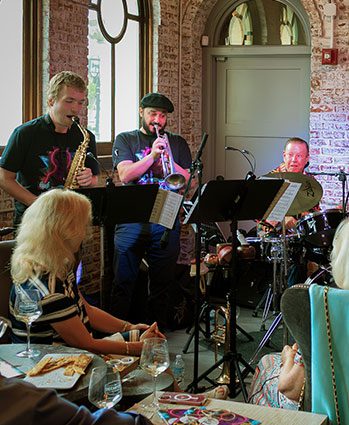 A small jazz band with saxophone, trumpet, and drums performs indoors for an audience seated at tables with drinks and food. Sunlight streams through tall windows behind the musicians.