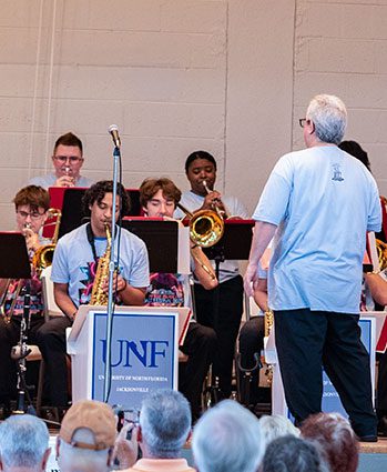 A conductor leads a group of young musicians playing brass and woodwind instruments onstage. UNF banners hang from their music stands. Audience members are seated in the foreground.