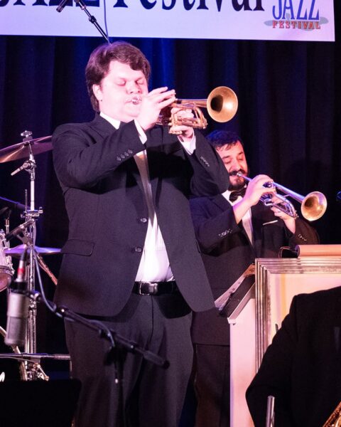 Two musicians in suits play trumpets on stage at a jazz festival, with a drum set partially visible on the left and a sign overhead reading JAZZ FESTIVAL..