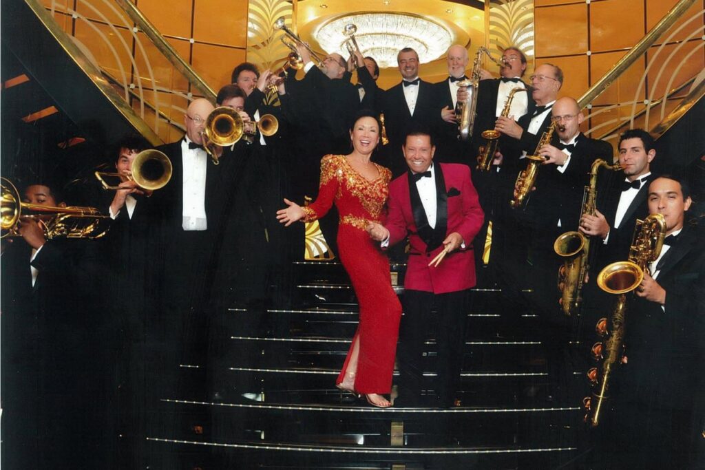 A woman in a red dress and a man in a red jacket pose on a staircase, surrounded by a jazz band in tuxedos playing brass and woodwind instruments.
