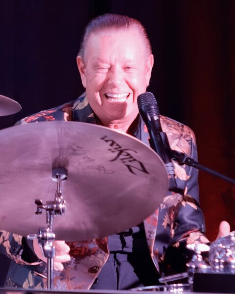 A smiling man in a floral jacket plays the drums and sings into a microphone, with a cymbal partially covering his face.