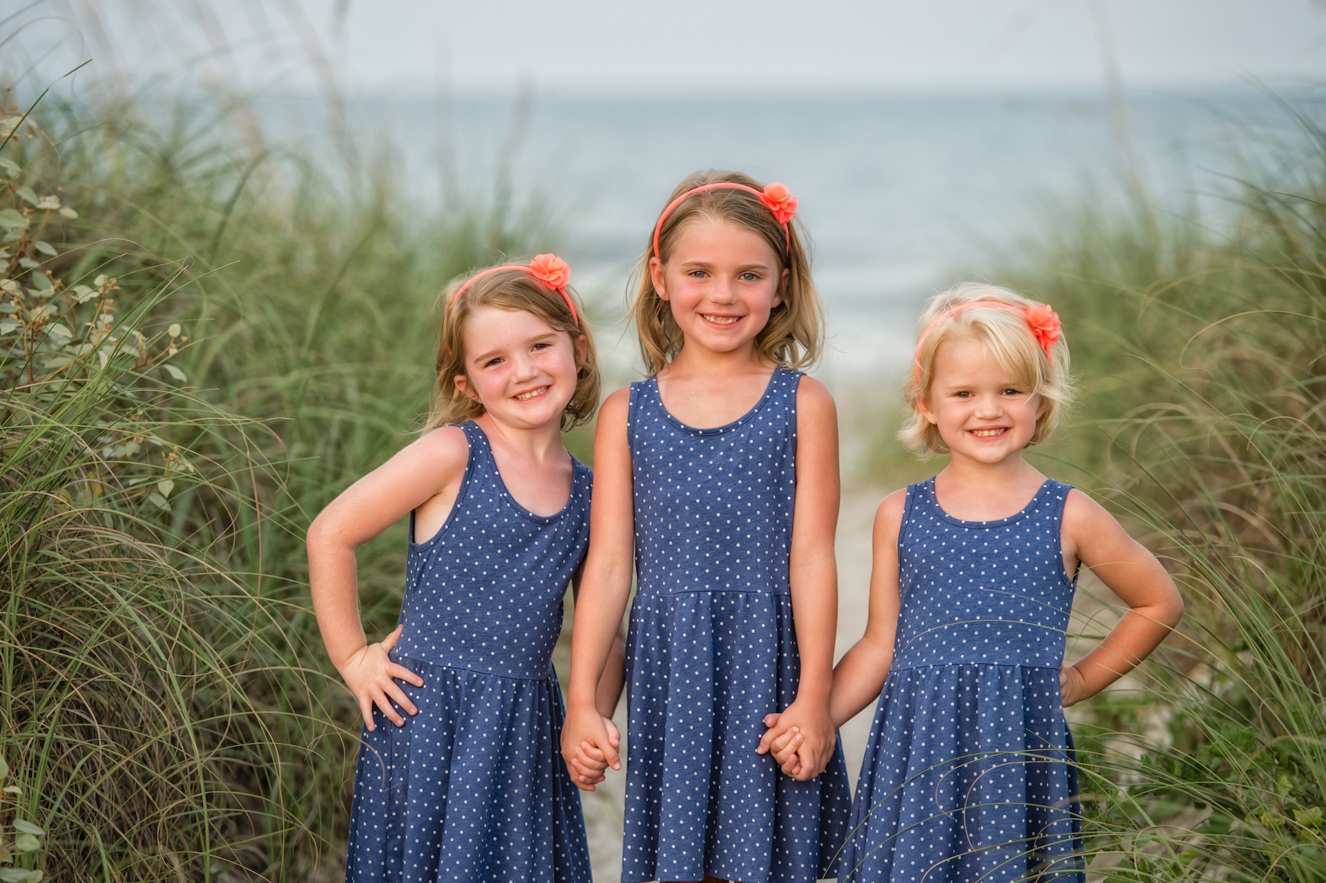 Three young girls in matching blue polka dot dresses and orange hair accessories stand smiling and holding hands on a sandy beach path lined with tall grass, with the ocean in the background.