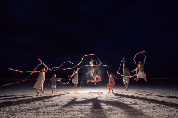 Seven people, including children and adults, jump in the air on a beach at night, each holding colorful ribbons. Their shadows stretch across the sand, and the scene is illuminated from behind.