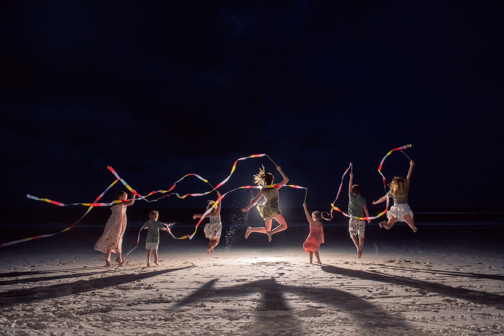 Seven people, including children and adults, jump in the air on a beach at night, each holding colorful ribbons. Their shadows stretch across the sand, and the scene is illuminated from behind.