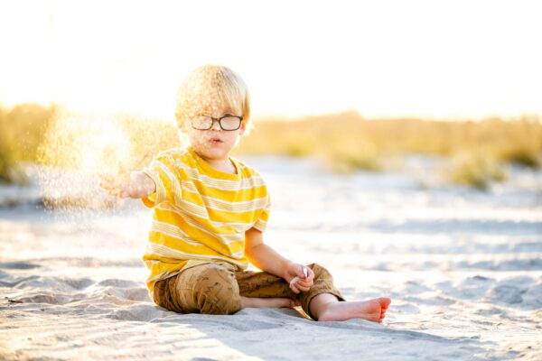 Young child wearing glasses and a yellow striped shirt sits on sandy ground, playfully tossing sand into the air on a sunny day at the beach. The background is bright and slightly blurred.