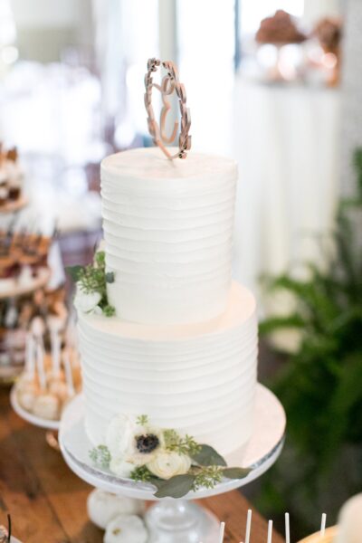 A two-tiered white wedding cake with textured frosting, decorated with white flowers and greenery, sits on a cake stand. The cake is topped with a metallic monogram topper.